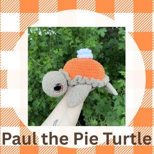 以下が含まれることがあります： オレンジ色のパイの形をした甲羅と白いホイップクリームが特徴の、かぎ針編みのカメのおもちゃ。カメは、茶色の手足、頭、目を持っています。「Paul the Pie Turtle」というテキストが下にあります。