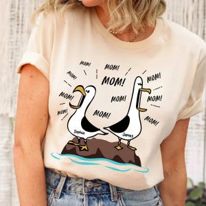 Puede incluir: Camiseta color crema con un diseño de dos gaviotas de dibujos animados sobre una roca, gritando "MOM!". Las gaviotas son negras y blancas con picos y patas amarillas. Los nombres "Sophia" y "James" están debajo.