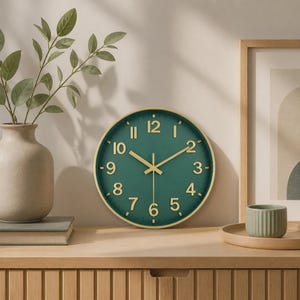 Könnte beinhalten: Eine runde Wanduhr mit dunkelgrünem Zifferblatt, goldenen Ziffern und goldenen Zeigern. Die Uhr verfügt über einen dünnen goldfarbenen Rahmen und ein schlichtes Design mit deutlichen Stundenmarkierungen sowie einem zentralen Uhrwerk.