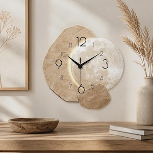 Può includere: Un orologio da parete con una cornice irregolare color beige effetto pietra, sovrapposta a un quadrante circolare illuminato con motivo lunare. Presenta lancette nere per ore e minuti, una lancetta dei secondi sottile e numeri arabi neri.