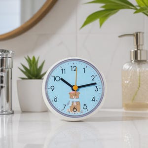 Könnte beinhalten: Eine runde analoge Uhr mit weißem Rahmen und hellblauem Zifferblatt. Die Uhr zeigt schwarze Zahlen von 1 bis 12, schwarze Stunden- und Minutenzeiger sowie einen gelben Sekundenzeiger. In der Mitte ist ein brauner Bär mit blauem Schal abgebildet.