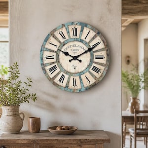 May include: A round wall clock with a distressed blue and cream finish, featuring black Roman numerals on a light background. The center dial displays the text "CAFEDELAGAR PARIS 1979" and "France" with black metal hour and minute hands.
