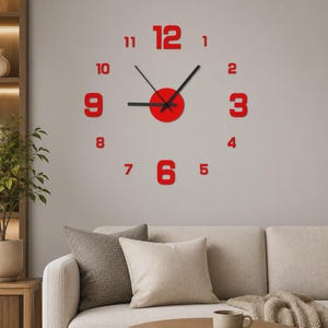 Könnte beinhalten: Eine Wanduhr mit großen, kräftigen roten Ziffern von 1 bis 12, die kreisförmig angeordnet sind. In der Mitte befindet sich ein rotes, kreisförmiges Element mit drei schwarzen Uhrzeigern für Stunden, Minuten und Sekunden.