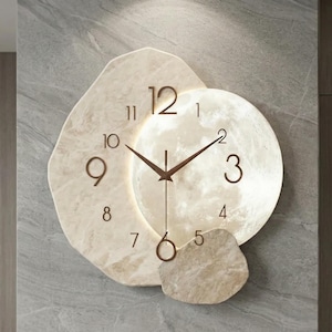 Pode incluir: Um relógio de parede moderno com um design único. O relógio apresenta uma moldura clara em forma de pedra irregular com um mostrador circular iluminado. Os ponteiros e os algarismos são de cor castanha escura, criando um contraste marcante.