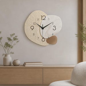 Peut inclure: Une horloge murale au design asymétrique composé de trois formes organiques superposées. La plus grande section présente une surface beige texturée, la section centrale un motif marbré blanc lisse et la plus petite est une pièce brune unie.