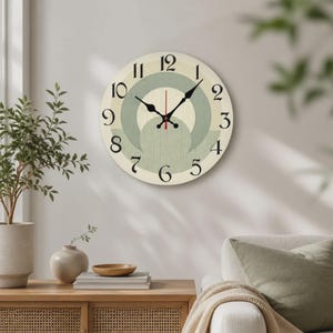 Könnte beinhalten: Eine runde Wanduhr mit cremefarbenem Zifferblatt und schwarzen arabischen Ziffern von 1 bis 12. Die Mitte zeigt überlappende, salbeigrüne kreisförmige geometrische Muster mit einer feinen vertikalen Textur. Schwarze Stunden- und Minutenzeiger.