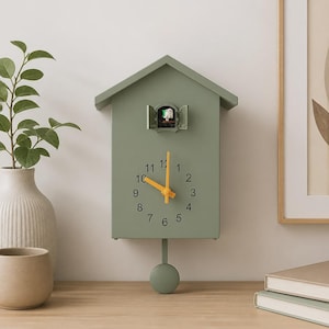 Pode incluir: Um relógio de parede verde-sálvia em forma de casa com telhado de duas águas. O mostrador apresenta números pretos de 1 a 12 e ponteiros amarelo-brilhantes. Uma pequena figura de pássaro branco está numa janela aberta e um pêndulo pende abaixo.
