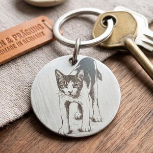 Puede incluir: Una etiqueta circular de metal plateado para llavero con una detallada ilustración grabada en negro de un gato de pie en posición frontal. La etiqueta está unida a una anilla de metal que también sostiene una llave de color latón.
