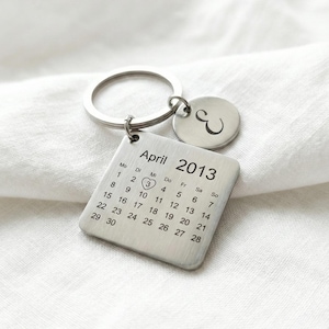 Peut inclure: Un porte-clés argenté avec un calendrier d'avril 2013. Le carré du calendrier indique le 3 avril avec un cœur. Un petit charme circulaire avec la lettre "E" est attaché. Le porte-clés est sur fond blanc.