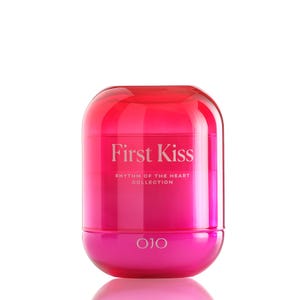May include: A cylindrical, two-toned container with a rounded top and base. The top is a translucent red, while the base is a solid pink. The text "First Kiss" is printed in white, along with "Rhythm of the Heart Collection" and the brand name "OJO".