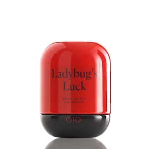 May include: A red and black container with the text "Ladybug's Luck" and "Spirit Animal Collection." The container has a rounded top and a black base with the OJO logo. The product is likely part of a beauty or wellness line.