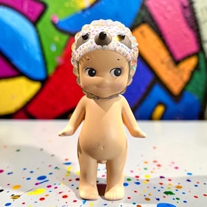 May include: A small, nude figurine with a smiling face and large eyes. The figurine wears a decorative head covering with a central brown design and iridescent accents. The background features colorful graffiti art and a speckled surface.