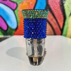 May include: A clear glass wallflower plug-in with a blue and gold jeweled top. The plug-in has a dark liquid inside. The background is a colorful graffiti design.