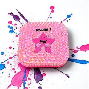 May include: A pink, square-shaped case covered in iridescent rhinestones. The case features a cartoon star graphic with the word "STARS!" above it. The background has pink, blue, and purple paint splatters.