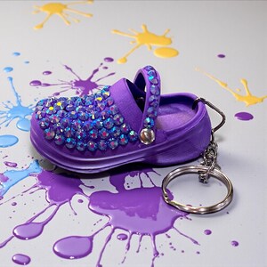 May include: A purple miniature shoe keychain, adorned with iridescent rhinestones on the front and strap. The keychain has a silver ring and clasp. The background features paint splatters in purple, yellow, and blue.