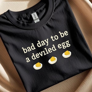 Può includere: T-shirt nero con la scritta bianca "bad day to be a deviled egg". Il design include tre illustrazioni di uova alla diavola. L'etichetta Gildan è visibile sul colletto.