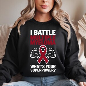 Peut inclure: Sweat-shirt noir avec le texte "I BATTLE MULTIPLE MYELOMA" en blanc et rouge. Un ruban rouge et un graphique de bras fléchissant sont en dessous du texte, avec les mots "WHAT'S YOUR SUPERPOWER?" en blanc.