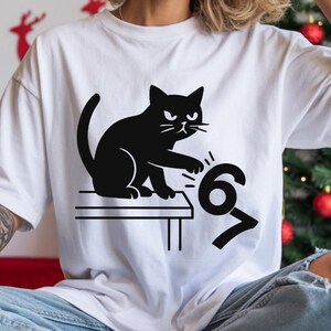 Pu&ograve; includere: T-shirt bianca con grafica di un gatto nero. Il gatto sembra colpire i numeri "67". Il design &egrave; minimalista e d'impatto, perfetto per l'uso quotidiano. Il disegno &egrave; stampato su una t-shirt bianca.