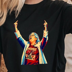 May include: A black short-sleeved t-shirt featuring a graphic of a person with blue hair and glasses, wearing a red shirt that says PRIMUS NO KINGS. The person has both arms raised with middle fingers extended in a stylised, colourful illustration.