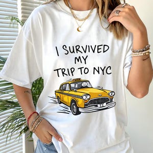 May include: A white short-sleeved t-shirt featuring the black text I SURVIVED MY TRIP TO NYC above a graphic illustration of a yellow taxi cab with a stick figure driver. The design is centered on the front of the garment.