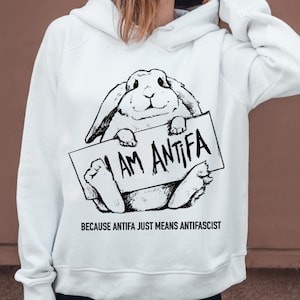 May include: White hoodie featuring a black and white illustration of a rabbit holding a sign that reads "I AM ANTIFA." Below the image, the text reads "BECAUSE ANTIFA JUST MEANS ANTIFASCIST."