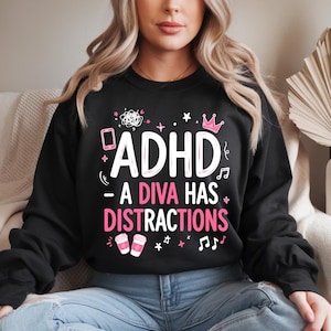 Può includere: Una felpa nera a maniche lunghe con testo grafico bianco e rosa che recita ADHD - A DIVA HAS DISTRACTIONS. Il design include illustrazioni di uno smartphone, una corona, tazze di caffè, note musicali, stelle e scarabocchi.