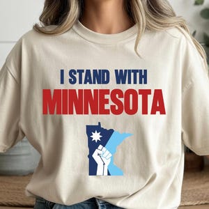 Peut inclure: T-shirt beige avec l'inscription "I STAND WITH MINNESOTA" en bleu et rouge. En dessous, un graphique de l'État du Minnesota avec un poing levé et une étoile.