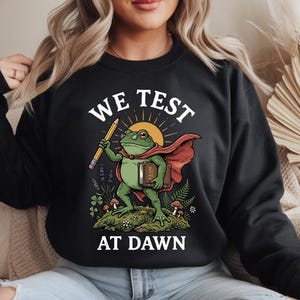 Peut inclure: Sweat-shirt noir avec un dessin d'une grenouille portant une cape et tenant un crayon et un livre. Le texte "WE TEST AT DAWN" est imprimé au-dessus et en dessous de l'illustration. La grenouille est sur un parterre d'herbe avec des champignons et des fleurs.