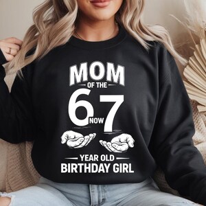 Peut inclure: Sweat-shirt noir avec texte blanc : "MOM OF THE 67 NOW YEAR OLD BIRTHDAY GIRL". Le design comprend un grand "67" et des mains stylisées. Un haut décontracté pour une fête d'anniversaire.