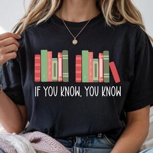 May include: Black t-shirt featuring a graphic of colorful books and the text "IF YOU KNOW, YOU KNOW" in white. The books are in shades of red, green, and pink. The shirt is a comfortable, casual style.