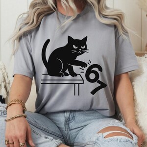 Pu&ograve; includere: T-shirt grigio chiaro con un disegno di gatto nero. Il gatto &egrave; seduto su un tavolo e sembra rovesciare i numeri 6 e 7. La maglietta &egrave; realizzata in un materiale morbido.