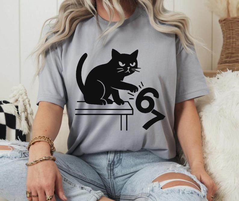 Pu&ograve; includere: T-shirt grigio chiaro con un disegno di gatto nero. Il gatto &egrave; seduto su un tavolo e sembra rovesciare i numeri 6 e 7. La maglietta &egrave; realizzata in un materiale morbido.