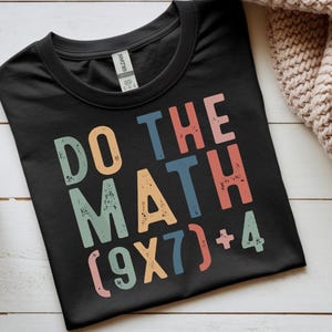 May include: Black t-shirt with the phrase "DO THE MATH" in colorful, distressed lettering. The equation (9X7)+4 is printed below the words. The shirt is made of a soft material.