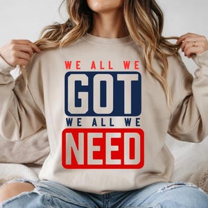 May include: A beige sweatshirt with the text "WE ALL WE GOT WE ALL WE NEED" in red and blue block letters. The sweatshirt is a neutral color and has long sleeves.