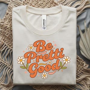 May include: Cream-colored t-shirt with the words "Be Pretti Good" in orange retro font, decorated with daisies and leaves. The shirt is folded neatly, with the brand tag visible at the neckline. A stylish and fun graphic tee.