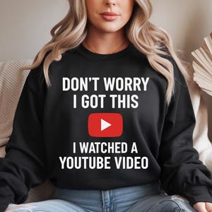 Pode incluir: Camisola preta com o texto "DON'T WORRY I GOT THIS" acima de um botão de reprodução do YouTube vermelho e o texto "I WATCHED A YOUTUBE VIDEO". A camisola é usada por uma pessoa.