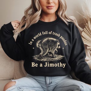 May include: A black long-sleeve sweatshirt featuring a graphic of a raccoon standing on leaves. The text above the raccoon reads "In a world full of trash pandas" and the text below reads "Be a Jimothy" in a light cream-colored font.