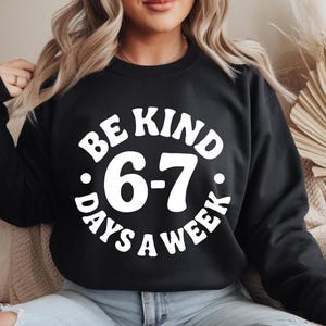 May include: Black sweatshirt with the white text "BE KIND 6-7 DAYS A WEEK" in a circular design. The sweatshirt is a crew neck style and is being worn by a person.