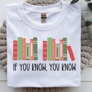 May include: White t-shirt with a graphic of colorful books in red, green, and pink. The text "IF YOU KNOW, YOU KNOW" is printed in black below the books. The shirt is folded on a textured surface.
