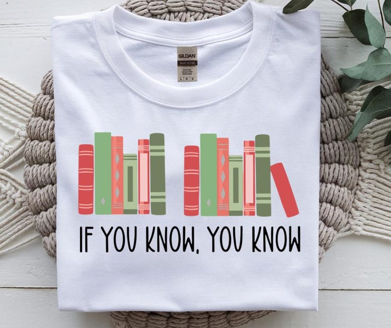 May include: White t-shirt with a graphic of colorful books in red, green, and pink. The text "IF YOU KNOW, YOU KNOW" is printed in black below the books. The shirt is folded on a textured surface.