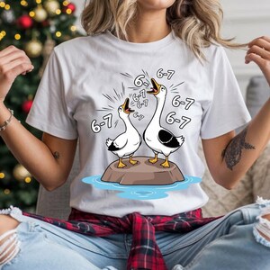 Peut inclure: T-shirt blanc avec une illustration de dessin animé de deux oies sur un rocher, avec le texte "6-7" au-dessus. Les oies sont noires et blanches avec des pieds jaunes. Le t-shirt est porté par une personne.