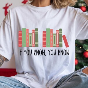 May include: White t-shirt featuring a graphic of colorful books in shades of red, green, and pink. The text "IF YOU KNOW, YOU KNOW" is printed below the books in black. The shirt is a casual, everyday wear item.