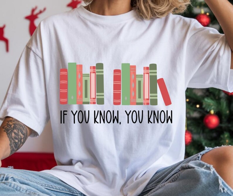 May include: White t-shirt featuring a graphic of colorful books in shades of red, green, and pink. The text "IF YOU KNOW, YOU KNOW" is printed below the books in black. The shirt is a casual, everyday wear item.