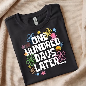 Peut inclure: T-shirt noir avec l'inscription blanche "ONE HUNDRED DAYS LATER...", entourée de fleurs de dessins animés colorées, d'une méduse rose et d'un hamburger. Le t-shirt est plié sur une surface beige.