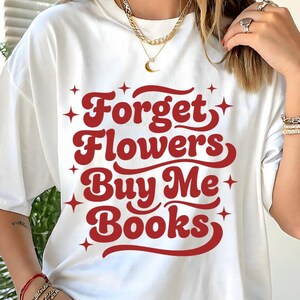 Peut inclure: T-shirt blanc avec l'inscription "Forget Flowers Buy Me Books" en rouge, dans un style rétro, ornée d'étoiles. Un vêtement décontracté pour tous les jours.
