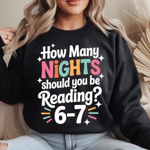 Peut inclure: Sweat-shirt noir avec l'inscription "How Many NIGHTS should you be Reading? 6-7" dans une typographie colorée et ludique. Le design comprend des étoiles et une police fantaisiste, parfait pour les lecteurs.