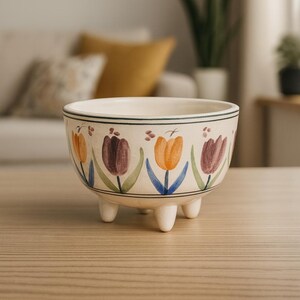 May include: A cream-colored ceramic bowl with three small feet. The bowl is decorated with a repeating pattern of orange and purple tulips with green leaves and blue stems. The rim has a black stripe.
