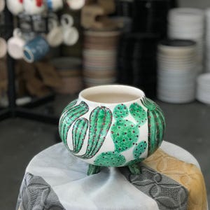 May include: A white ceramic pot with a green cactus design and red accents. The pot has a rounded shape with three small feet. The interior is white, and the pot is displayed on a patterned surface.