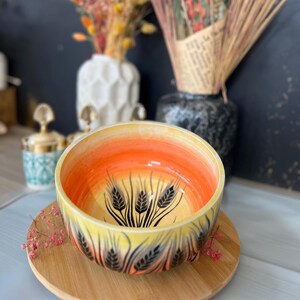 May include: A ceramic bowl with a wheat design, featuring a gradient of yellow, orange, and black. The bowl sits on a round wooden plate, with dried flowers in the background.
