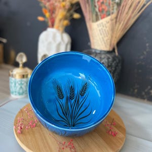May include: A vibrant blue ceramic bowl with a wheat stalk design. The bowl is set on a wooden plate, with dried flowers in the background. The wheat design is black and adds a rustic touch to the bowl.
